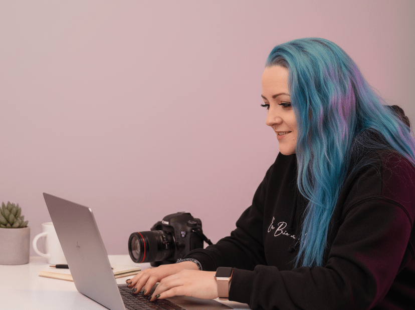 Woman with vivid blue‑green hair types on a silver laptop at a white desk, with a camera, plant, and mug nearby.